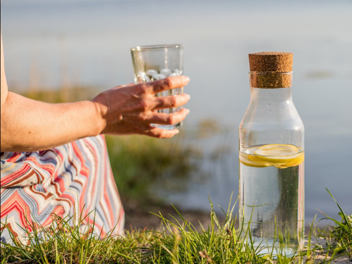 A woman drinks water with lemon from a glass. A glass carafe of water with lemon stands next to her.